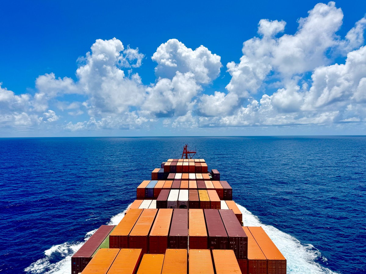 Forward view from container ship deck showing stacked cargo containers and open ocean with blue sky and clouds, symbolizing global trade and maritime logistics